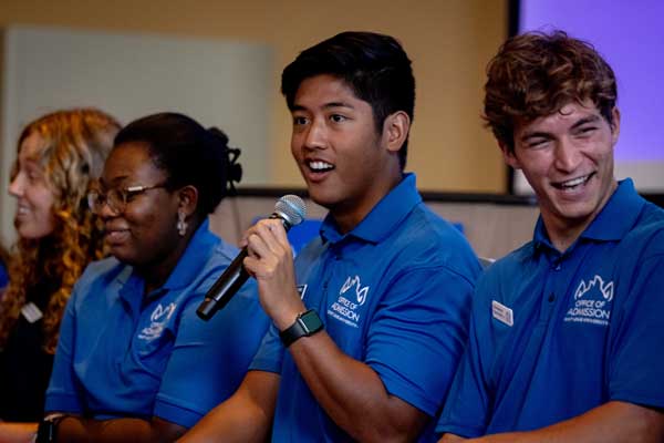 A student speaks into a microphone. Two students are seated next to him, smiling.