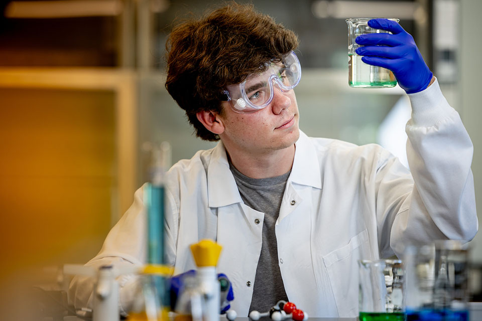 A student in lab protective hear holds a beaker up to the light 