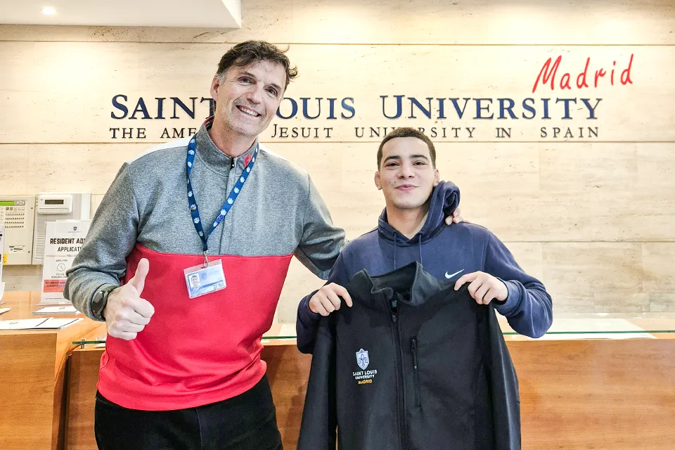 Sports director César Rioja (left) and junior Jack Montoya (right) pose in front of San Ignacio Hall’s reception when Montoya received his SLU-Madrid sweatshirt. Man to the left gives a thumbs up and smiles at camera and student to his right looks on at camera while holding a sweater with the university logo. There is the university logo on the wall in the background behind a reception desk.