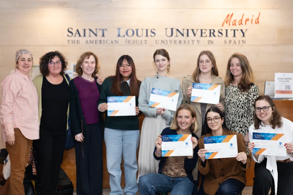 SLU-Madid faculty pose alongside student presenters who received participation certificates in the San Ignacio Hall lobby. Three women standing look onward at camera next to three students posing with certificates standing in the same row. There are three other students posing with the same certificate who are kneeling with a woman standing behind them. There is a logo on the wall in the background.