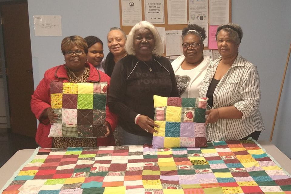 A group of older adults in a Circle of Friends group pose in front of a large patchwork quilt displayed on the table before them.
