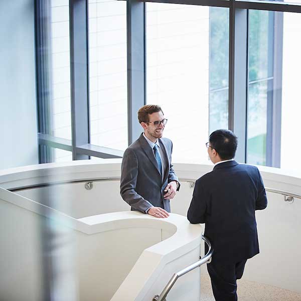 Two students where suits, ties, and glasses laugh and enjoy conversation on a large, circular, and modern staircase in front of large windows. 